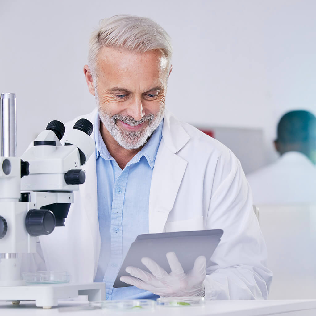 Ein Professor liest etwas auf seinem Tablet und lächelt. Neben ihm steht ein Mikroskop.